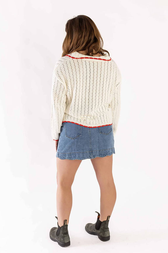 Woman wearing Weaving Color white cardigan knit with red trim paired with denim skirt and black boots
