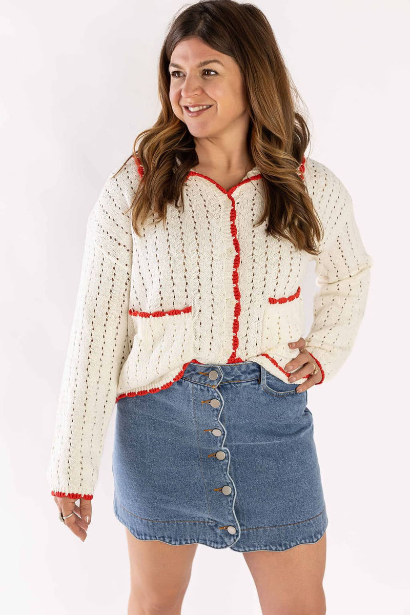 Woman wearing Weaving Color white cardigan knit with red trim and front pockets paired with a button-up denim skirt
