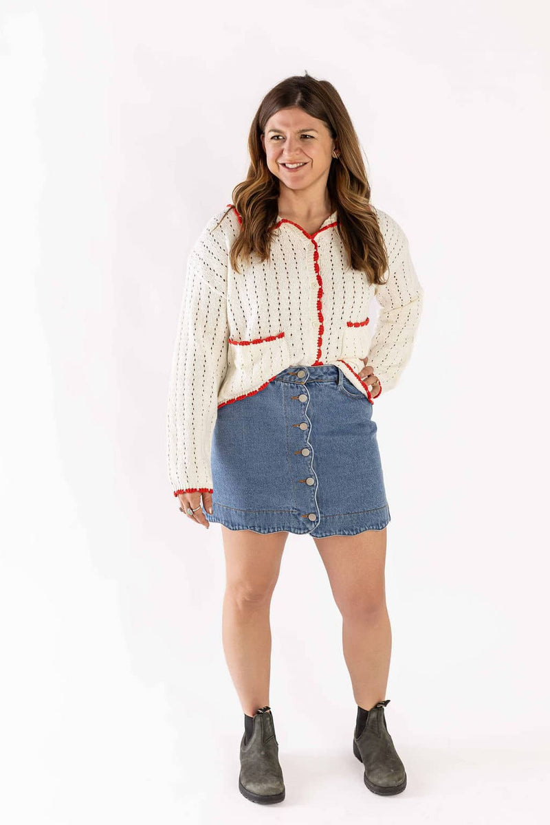 Woman wearing white cardigan knit with red trim paired with a button-front denim skirt and ankle boots