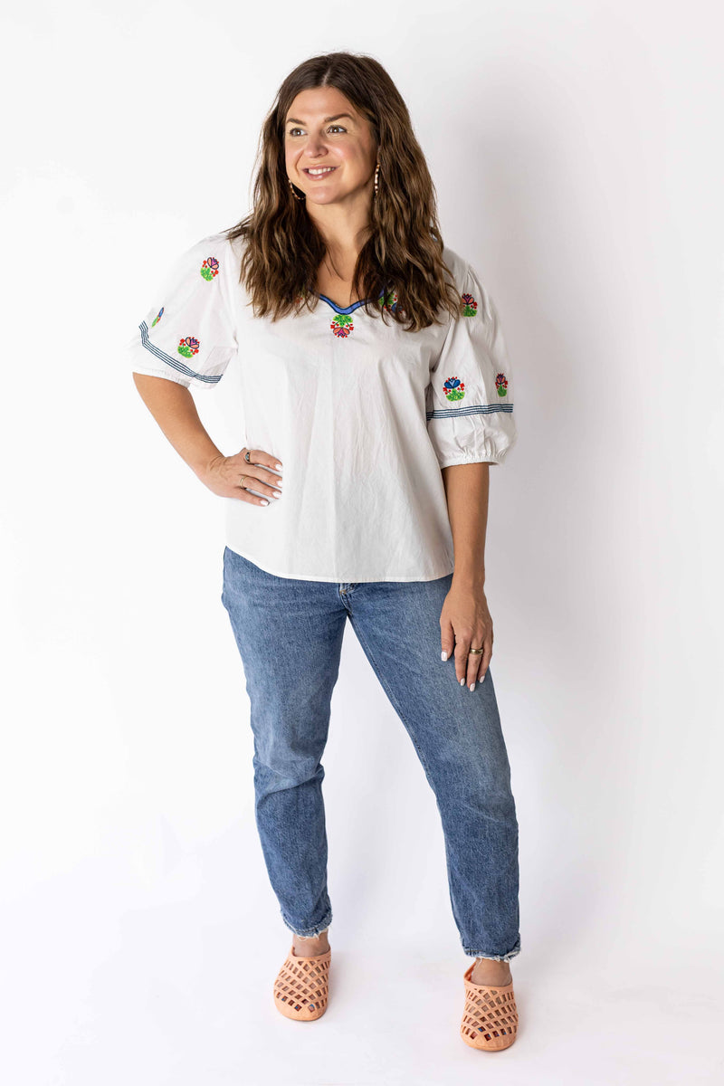 Woman wearing white embroidered peasant top with colorful floral designs paired with blue jeans