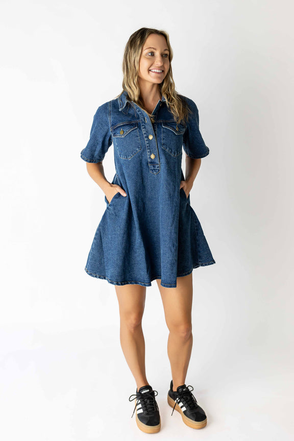 Woman wearing a medium wash denim dress with short sleeves and front pockets paired with black sneakers looking forward