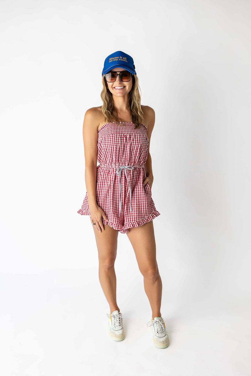 Woman wearing a red checkered tube romper with ruffle hem and drawstring waist paired with sneakers and a blue cap