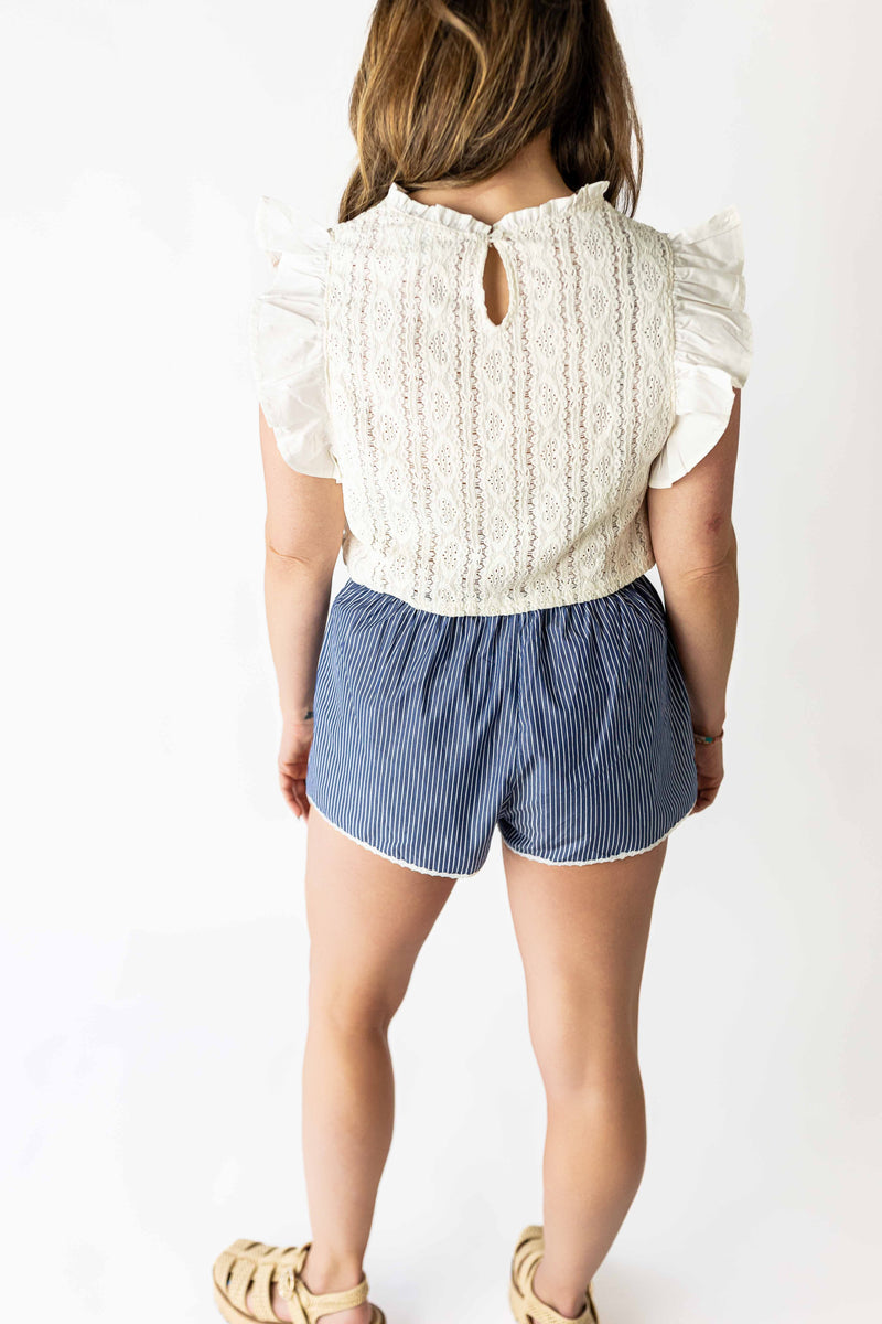 Back view of woman wearing white ruffle sleeve lace top with keyhole detail paired with blue striped shorts