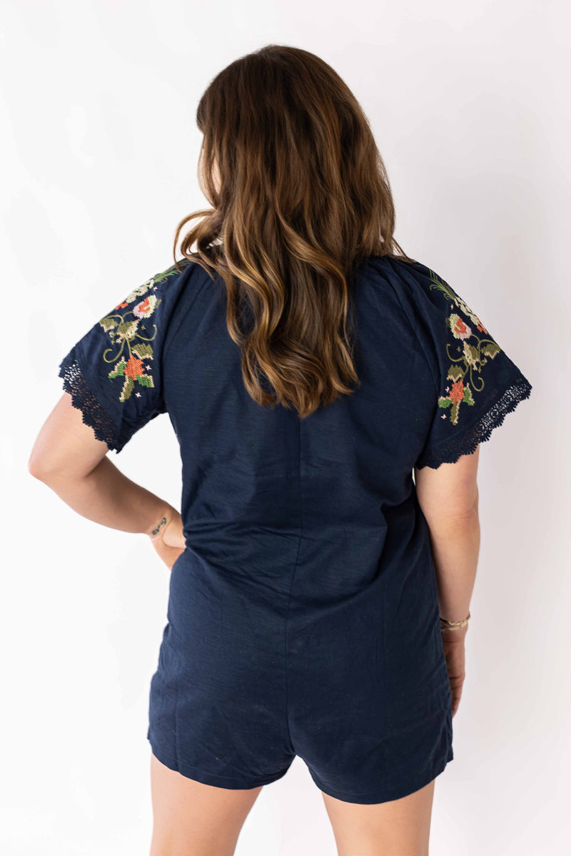 Back view of woman wearing navy embroidered bloom romper with floral sleeve details by Weaving Color