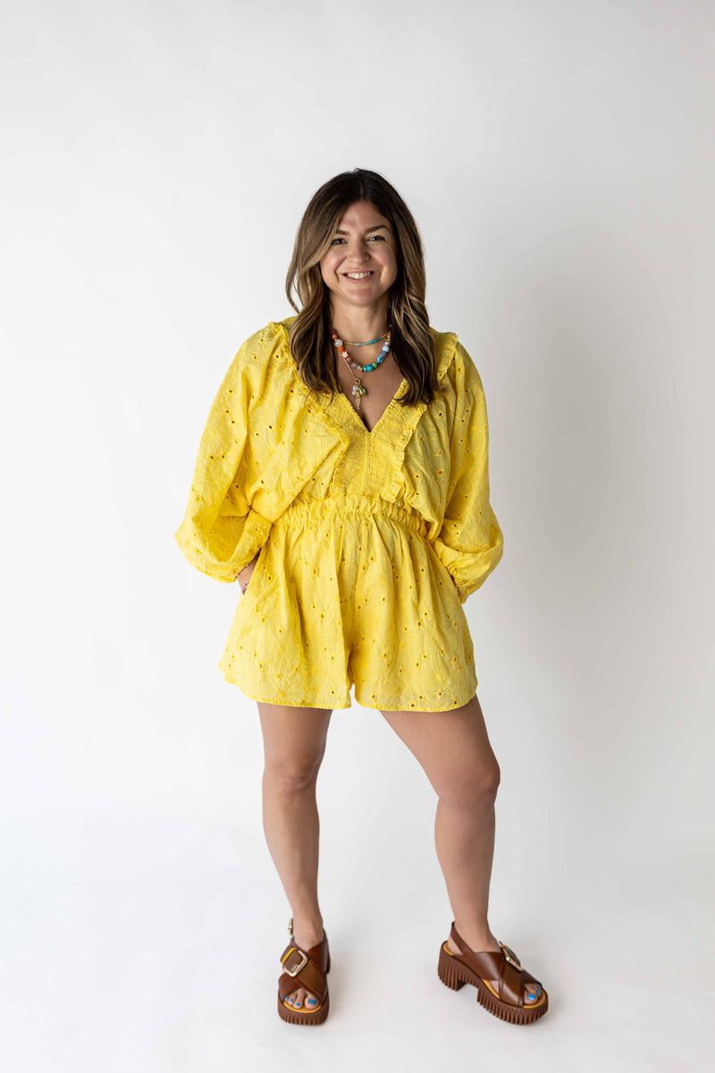 Smiling woman wearing a yellow eyelet romper with long sleeves and a cinched waist paired with brown sandals